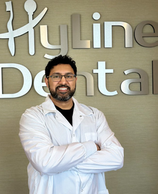 The image shows a man standing in front of a sign that reads HYLINE DENTAL. He is wearing a white lab coat and has his arms crossed.