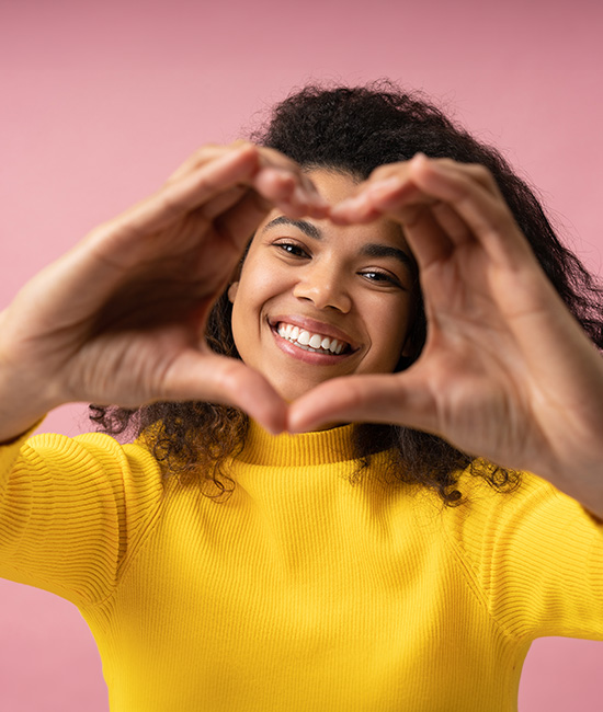 A woman is holding her hands together in a heart shape, smiling brightly at the camera. She has dark hair and is wearing a yellow top, standing against a pink background.