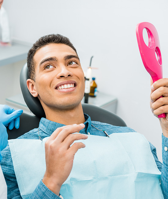 The image shows a man sitting in a dental chair, smiling at the camera, with a dental hygienist or dentist standing behind him holding a pink oral care device.