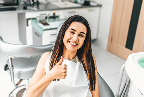 A woman with a thumbs-up gesture is seated in a dental chair, smiling at the camera.