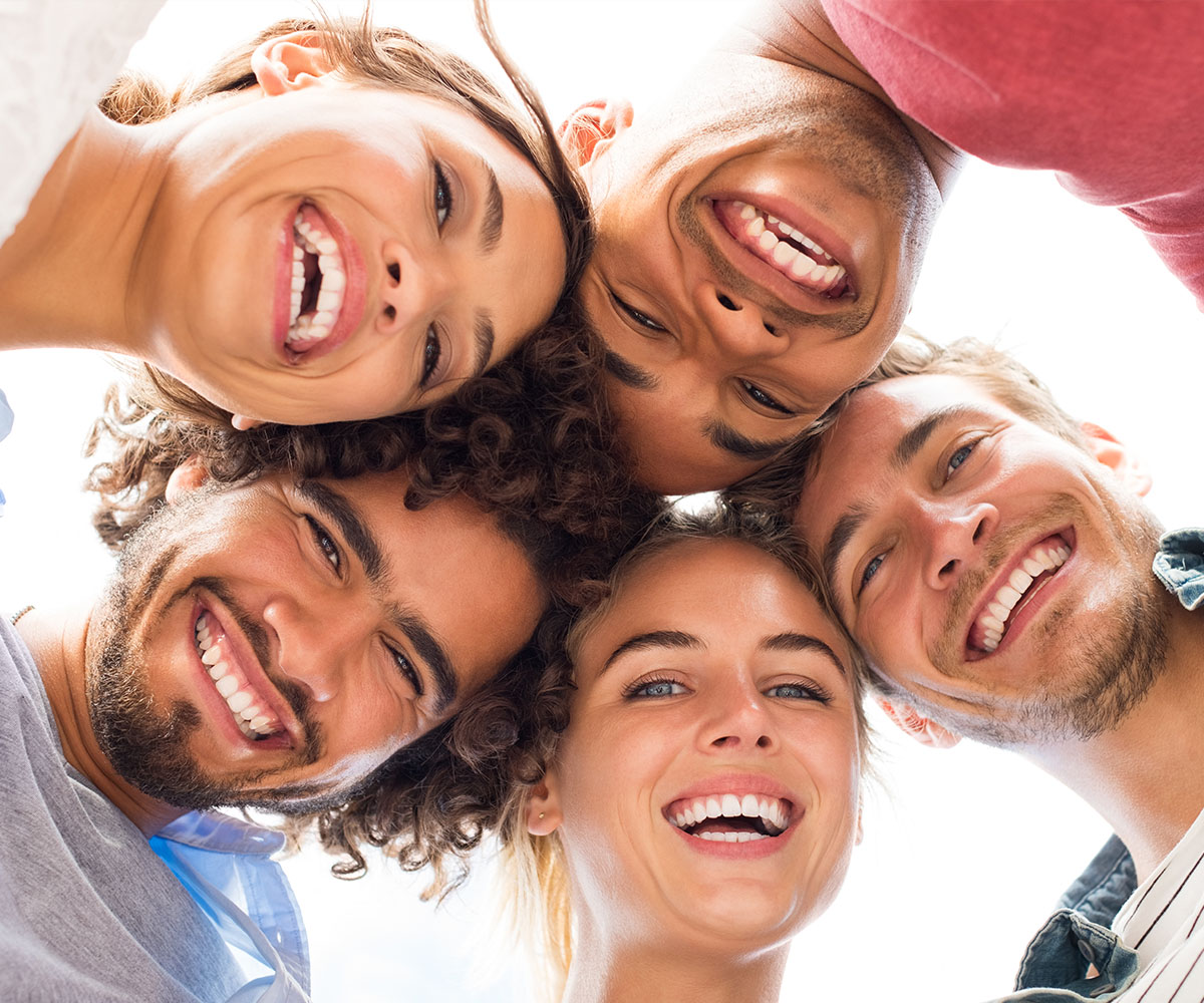 A group of six young adults, three male and three female, are smiling at the camera with their arms around each other.